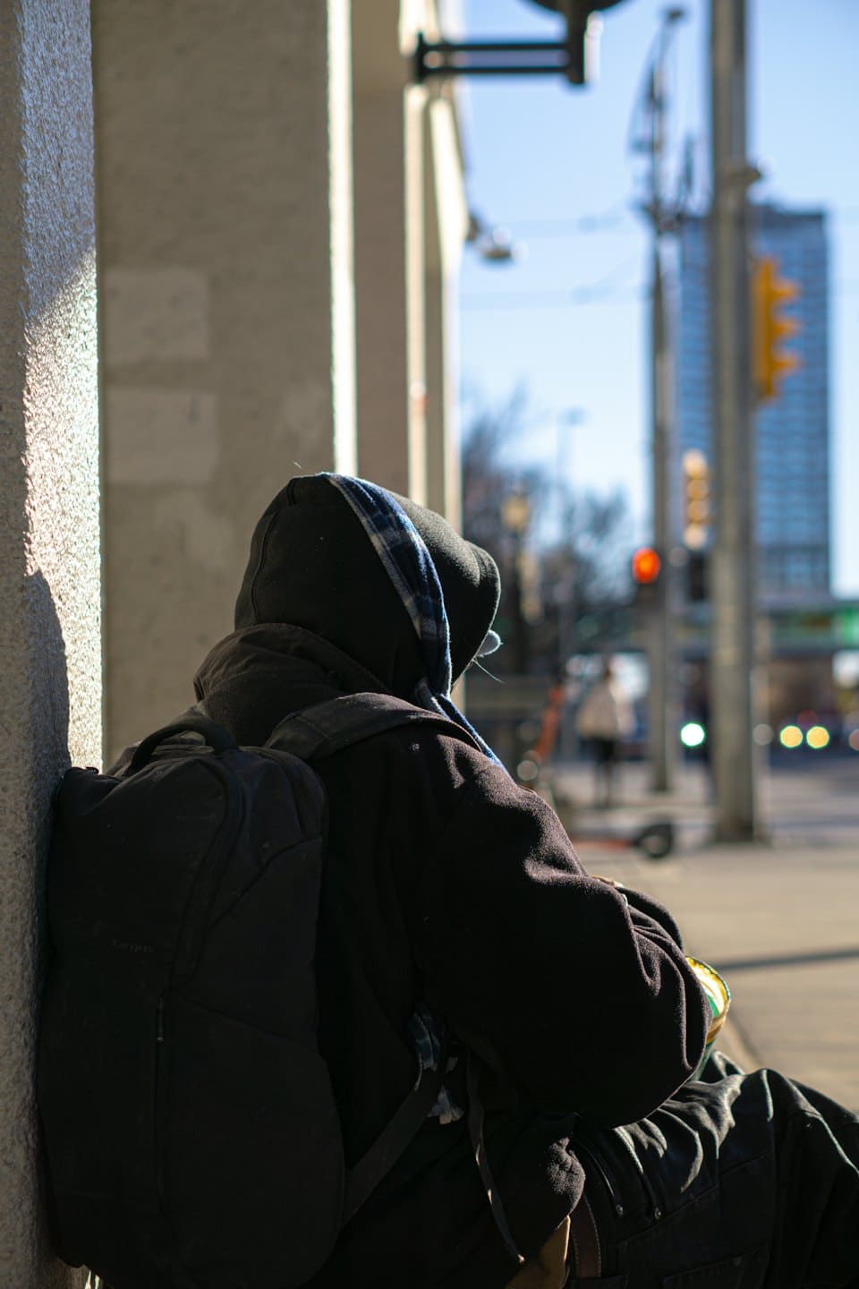 Eine Person mit schwarzer Kapuze und Rucksack lehnt an einer Säule.