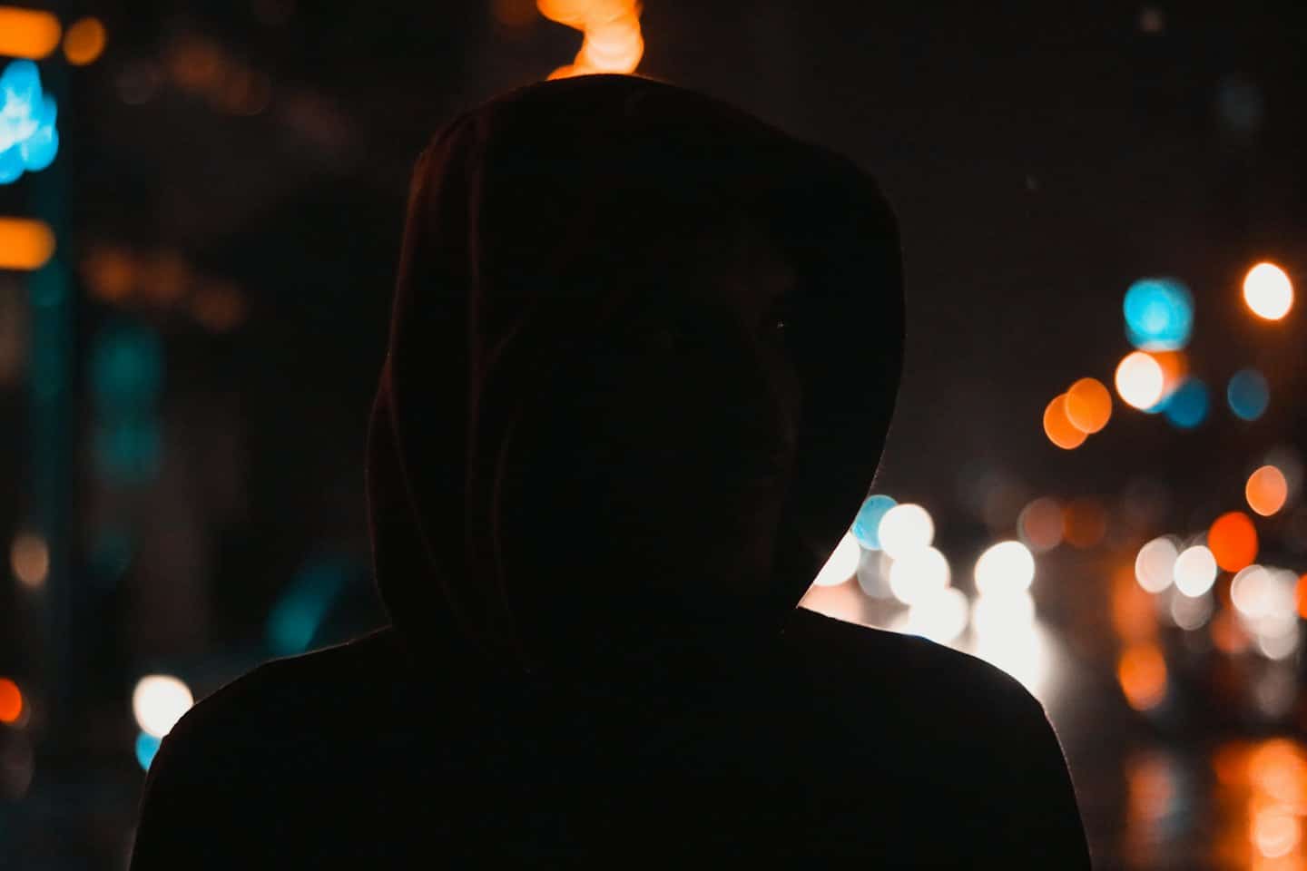 Kapuzenfigur vor unscharfen Stadtlichtern bei Nacht.